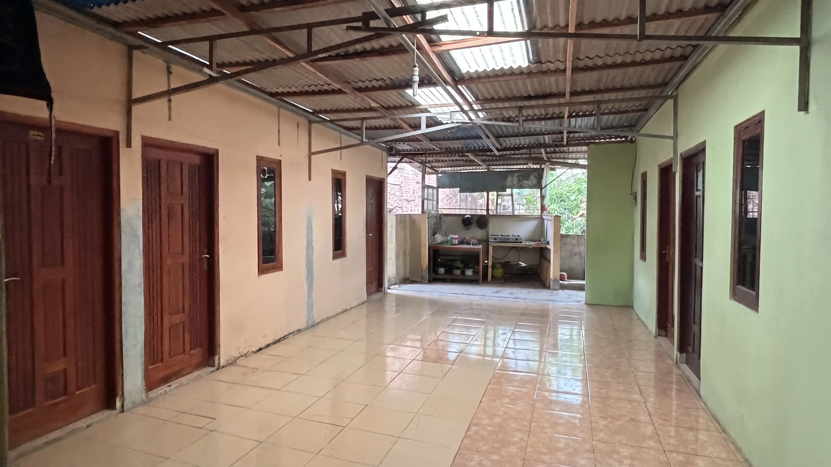 Lorong kamar berlantai keramik dengan deretan pintu kamar, dapur bersama terlihat di ujung